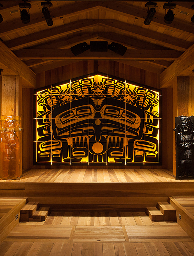 An illuminated formline clan house screen in a wood-paneled hall.