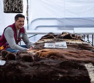 A Native artist sits behind a market table displaying furs
