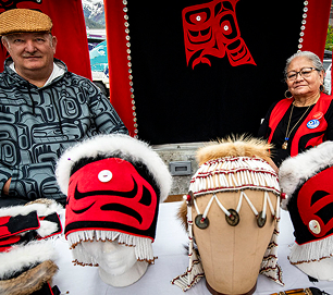 Two Native artists display red, black, and white regalia and woven hats