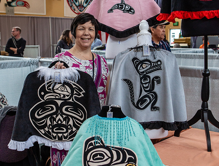 A smiling Native artist displays garments with Northwest Coast designs