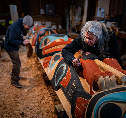 Two artists carving and painting a monumental cedar Kootéeyaa