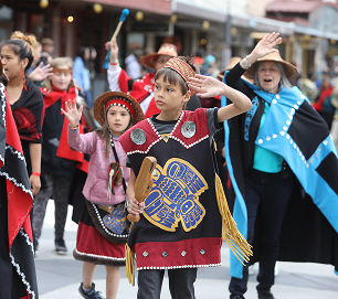 Young parade participants in colorful regalia wave to spectators
