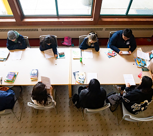 Students draw at tables beside large classroom windows