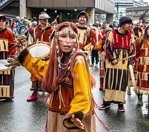 A dancer in golden regalia performs at the front of a drum group