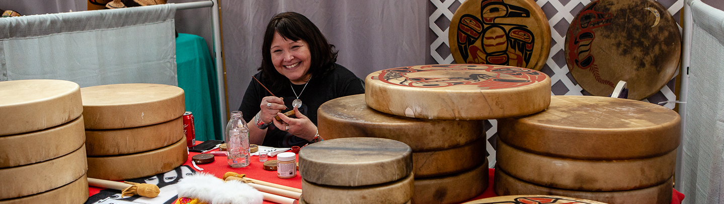 An Alaska Native artist painting a hand drum at an art market table