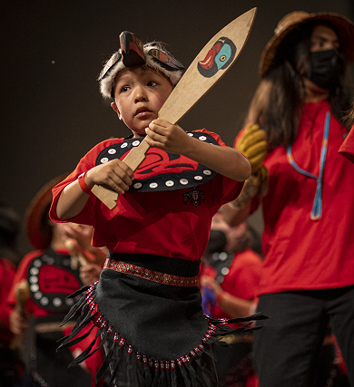 A young dancer in regalia holds a carved dance paddle