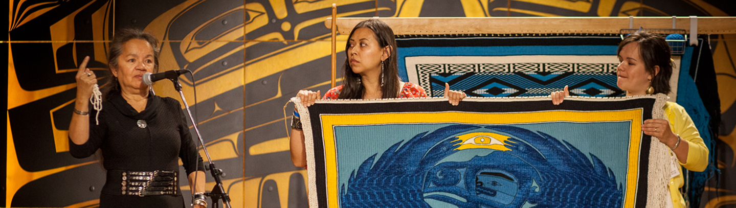 Three women presenting a blue woven robe during a Northwest Coast arts lecture