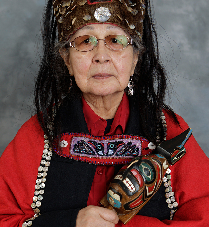 Ruth Demmert wearing regalia and holding a carved Raven rattle