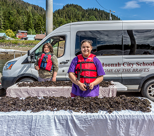 Students stand behind a table covered with black seaweed near a Hoonah City Schools van