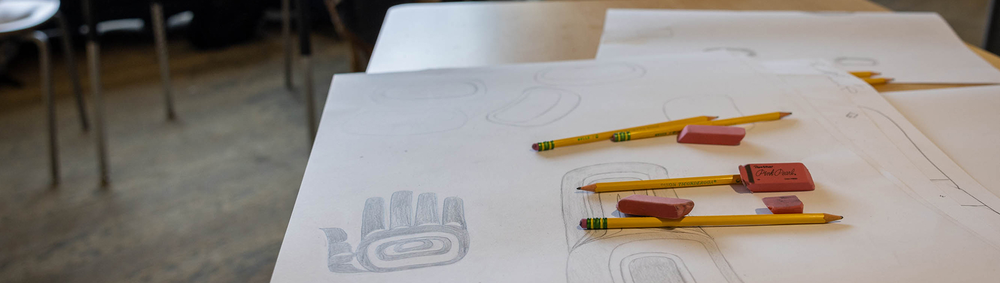 Formline drawings, pencils, and erasers arranged on a classroom table