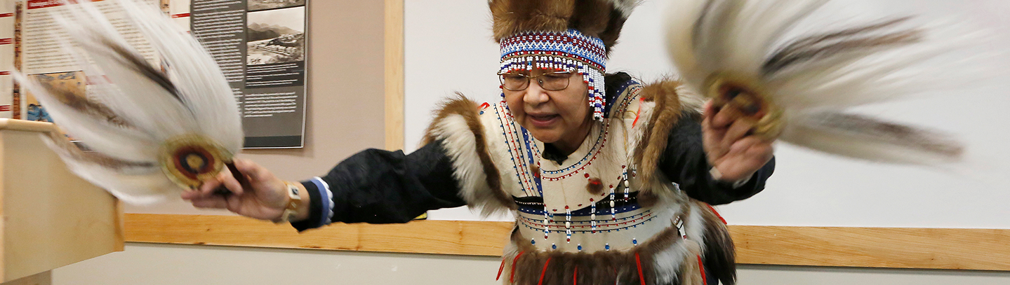 A cultural presenter in regalia holding feather fans during a lecture