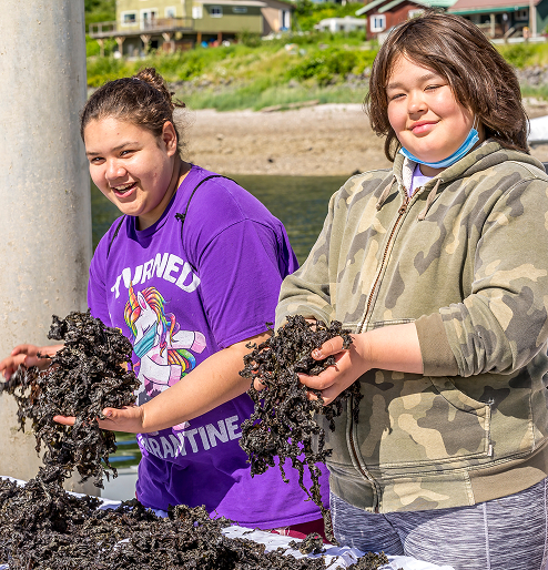 Two young people smile while holding freshly harvested black seaweed on the waterfront